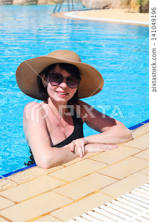 Woman in sunglasses and wide-brimmed hat swimming leisurely in a sunny pool Woman in sunglasses and wide-brimmed hat swimming leisurely in a sunny pool 141049196