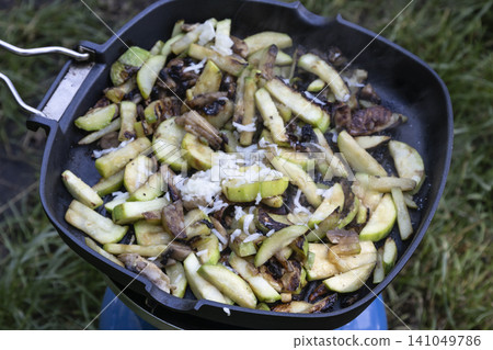 Close-up of sliced zucchini and mushrooms sizzling and steaming in a black frying pan on a portable blue camping stove, cooked outdoors over a grass background during a picnic or hike. Close-up of sliced zucchini and mushrooms sizzling and steaming in a black frying pan on a portable blue camping stove, cooked outdoors over a grass background during a picnic or hike. 141049786