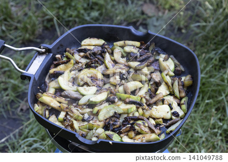 Close-up of sliced zucchini and mushrooms sizzling and steaming in a black frying pan on a portable blue camping stove, cooked outdoors over a grass background during a picnic or hike. Close-up of sliced zucchini and mushrooms sizzling and steaming in a black frying pan on a portable blue camping stove, cooked outdoors over a grass background during a picnic or hike. 141049788