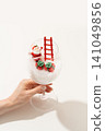 Creative Christmas concept photography of a hand holding a snow-filled wine glass with festive miniature decorations on a clean white studio background. 141049856
