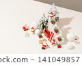 Overhead Christmas flat lay with snow-covered tree, ornament balls, and festive figurines arranged on a white background with strong directional shadow for seasonal advertising use. 141049857