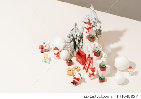 Overhead Christmas flat lay with snow-covered tree, ornament balls, and festive figurines arranged on a white background with strong directional shadow for seasonal advertising use. 141049857