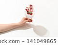 Female hand holding a clear wine glass with a miniature Santa Claus climbing a small ladder and gift boxes 141049865