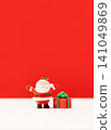 Miniature Santa Claus figurine peeking around a white corner prop next to a red gift box 141049869