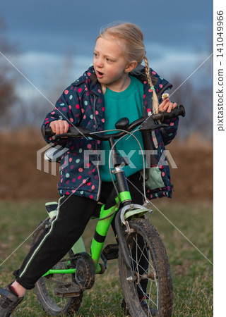 Young blonde girl with a braid sitting on a green bicycle, wearing a colorful polka dot jacket and looking with a surprised expression across a grassy field under a cloudy sky Young blonde girl with a braid sitting on a green bicycle, wearing a colorful polka dot jacket and looking with a surprised expression across a grassy field under a cloudy sky 141049966