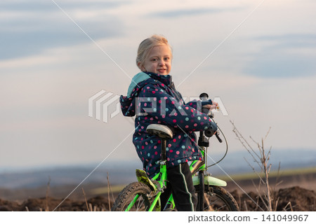 Young blonde girl wearing a polka dot jacket and black pants, smiling while standing with her bicycle on an unpaved outdoor path, looking directly at the viewer Young blonde girl wearing a polka dot jacket and black pants, smiling while standing with her bicycle on an unpaved outdoor path, looking directly at the viewer 141049967