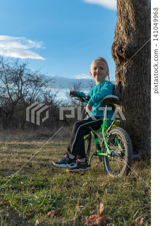 Young child with a happy expression sitting on a bicycle leaning against a tree trunk, enjoying a moment of outdoor play in a natural setting on a sunny day Young child with a happy expression sitting on a bicycle leaning against a tree trunk, enjoying a moment of outdoor play in a natural setting on a sunny day 141049968