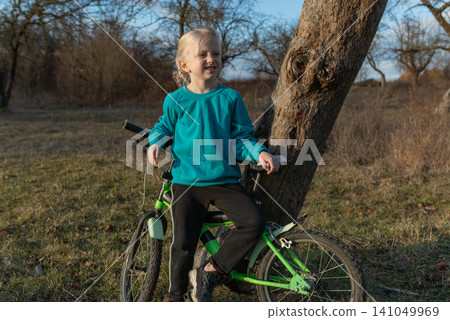 Blonde girl leaning against a tree with a green bicycle in a sunny grassy field, smiling and enjoying outdoor play, childhood freedom and simple nature moments Blonde girl leaning against a tree with a green bicycle in a sunny grassy field, smiling and enjoying outdoor play, childhood freedom and simple nature moments 141049969