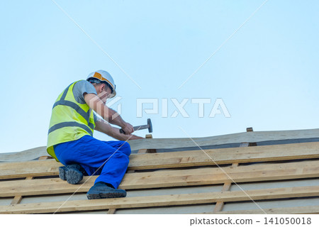 Construction worker in hard hat and safety vest nailing wooden battens for roof underlayment on a new house frame, demonstrating skilled carpentry and on site craftsmanship Construction worker in hard hat and safety vest nailing wooden battens for roof underlayment on a new house frame, demonstrating skilled carpentry and on site craftsmanship 141050018