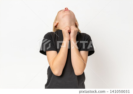 Young child with blonde hair wearing a t shirt, looking upwards with hands clasped in a gesture of hope or seeking help, standing on a white background 141050031