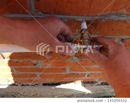 A close-up shot shows male hands connecting white plumbing pipes to a brass tee fitting. The plumbing assembly is being installed directly against a textured wall of red brick and cement. A close-up shot shows male hands connecting white plumbing pipes to a brass tee fitting. The plumbing assembly is being installed directly against a textured wall of red brick and cement. 141050332