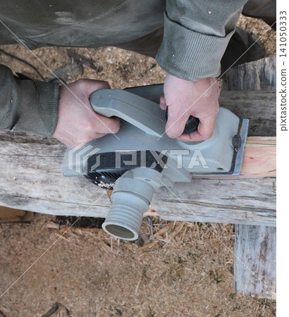 A pair of male hands confidently guides a grey electric planer down the length of an old log, removing the top layer of wood; high-angle view. Fresh sawdust and wood shavings lie scattered around. A pair of male hands confidently guides a grey electric planer down the length of an old log, removing the top layer of wood; high-angle view. Fresh sawdust and wood shavings lie scattered around. 141050333