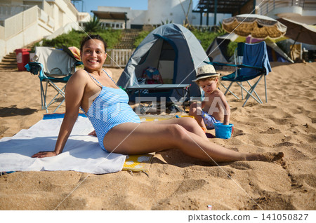 Pregnant woman and toddler playing on sandy beach near tents during a sunny family vacation Pregnant woman and toddler playing on sandy beach near tents during a sunny family vacation 141050827