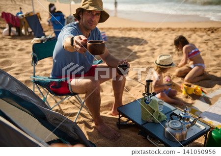 Man offering coffee on a family beach camping trip with children playing in the sand Man offering coffee on a family beach camping trip with children playing in the sand 141050956