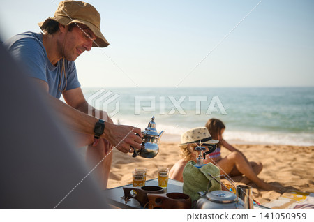 Father pouring tea for family on sandy beach during relaxed summer picnic by the sea 141050959