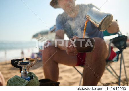 Man pouring hot coffee into wooden cup at the beach during a relaxed camping morning Man pouring hot coffee into wooden cup at the beach during a relaxed camping morning 141050960