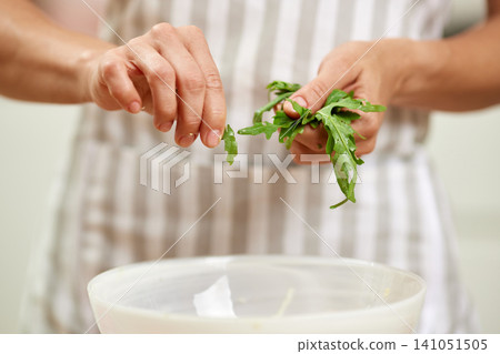 Person's hands tearing fresh arugula leaves into a bowl, preparing a healthy and fresh salad in the kitchen 141051505