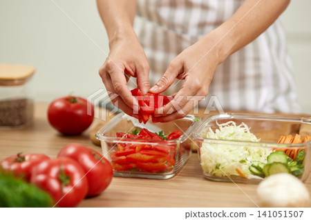 Woman's hands placing chopped fresh tomatoes into a glass food container, preparing healthy meal preps on a wooden kitchen counter 141051507