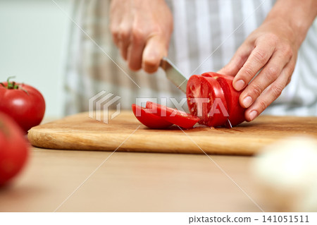 Hands slicing fresh red tomatoes on a wooden cutting board, preparing food in a kitchen for healthy cooking and eating Hands slicing fresh red tomatoes on a wooden cutting board, preparing food in a kitchen for healthy cooking and eating 141051511