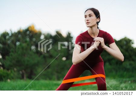 Woman in red outfit exercises outdoors using resistance band in the park Woman in red outfit exercises outdoors using resistance band in the park 141052017