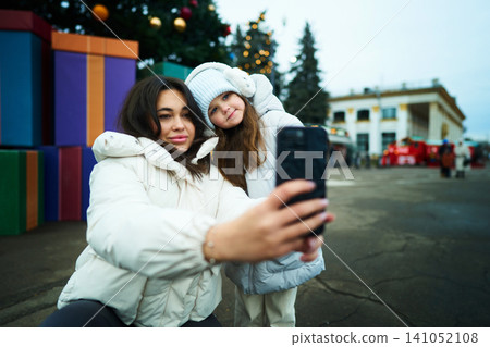 Two girls take a selfie in front of festive decorations at a winter market Two girls take a selfie in front of festive decorations at a winter market 141052108