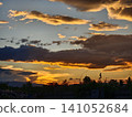 A dramatic sunset landscape with vibrant golden and orange clouds in a deep blue sky. Black tree silhouettes on the horizon create a peaceful, yet powerful natural background scene. 141052684