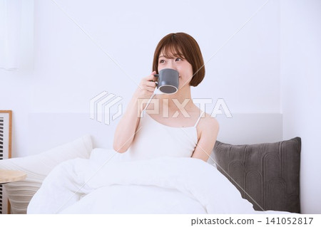 清晨,一位年輕女子躺在床上,手裡拿著杯子喝咖啡。 清晨,一位年輕女子躺在床上,手裡拿著杯子喝咖啡。 141052817