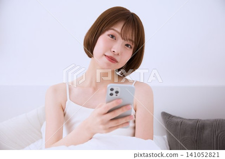 一個年輕的女人在床上操作智能手機 一個年輕的女人在床上操作智能手機 141052821