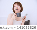 A young woman smiles, relaxing with her smartphone and mug in her hands. 141052826