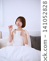 A smiling young woman brushing her teeth in bed in the morning, holding a coffee cup. 141052828