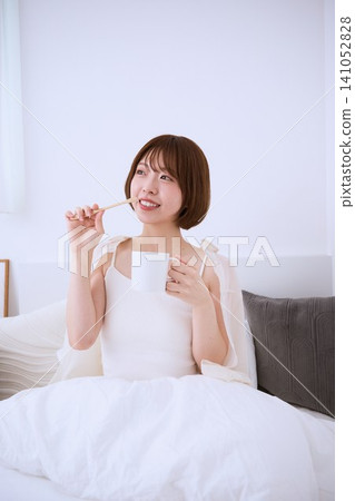 一位微笑的年輕女子早上在床上刷牙,手裡拿著一杯咖啡。 一位微笑的年輕女子早上在床上刷牙,手裡拿著一杯咖啡。 141052828