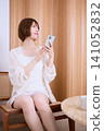 A young woman in white clothes is sitting in a chair and operating her smartphone. 141052832