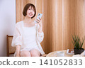 A young woman in a white dress smiles while holding her smartphone in a cafe. 141052833