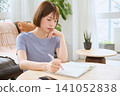 A young woman is thinking while writing in her notebook in the living room. 141052838