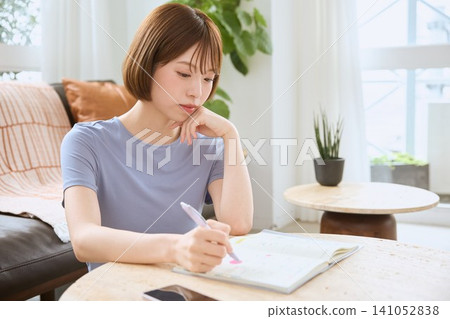 一位年輕女子在客廳裡一邊在筆記本上寫字一邊思考。 141052838