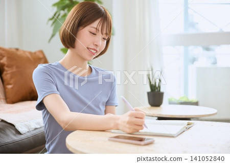 一位留著波波頭、微笑的年輕女子正在客廳的筆記本上寫東西。 一位留著波波頭、微笑的年輕女子正在客廳的筆記本上寫東西。 141052840