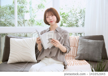 A young woman with bobbed hair, smiling, is sitting on a sofa reading a book. A young woman with bobbed hair, smiling, is sitting on a sofa reading a book. 141052843