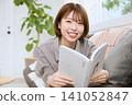 A young woman smiles while reading a book on a sofa. 141052847