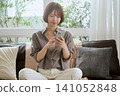 A smiling young woman using her smartphone on the living room sofa. 141052848