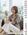 A young woman sitting on a sofa, looking at her smartphone and laughing. 141052850