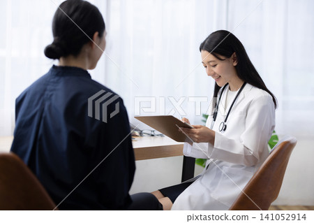 A young Japanese female doctor writing in a patient's medical record during an examination. A young Japanese female doctor writing in a patient's medical record during an examination. 141052914