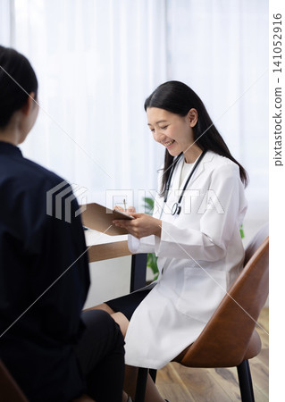 A young Japanese female doctor writing in a patient's medical record during an examination. A young Japanese female doctor writing in a patient's medical record during an examination. 141052916