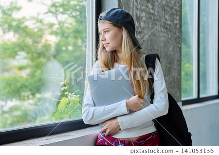 Pensive teenage girl student holding laptop and standing by large window Pensive teenage girl student holding laptop and standing by large window 141053136