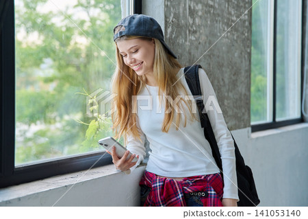 Smiling 16-year-old teenage girl using smartphone by the window Smiling 16-year-old teenage girl using smartphone by the window 141053140