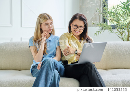 Skeptical teenage daughter sitting with smiling mother looking at laptop Skeptical teenage daughter sitting with smiling mother looking at laptop 141053233