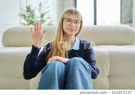 Smiling teenage girl in glasses waving hand during online video call. Smiling teenage girl in glasses waving hand during online video call. 141053237