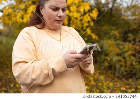 woman texting on phone in outdoor autumnal scene with trees and foliage, close-up, slightly low angle, focused, beige sweatshirt, plastic phone case, natural light, clean background. woman texting on phone in outdoor autumnal scene with trees and foliage, close-up, slightly low angle, focused, beige sweatshirt, plastic phone case, natural light, clean background. 141053744