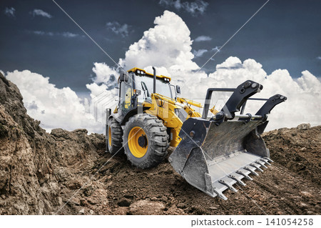 A universal excavator loader is working at a construction site. The loader is digging into dirt while clouds fill the sky above 141054258