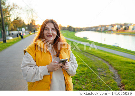 woman holding phone on outdoor pathway near water body, casual and relaxed scene with concrete and grass materials, yellow and green colors, modern and bright mood. woman holding phone on outdoor pathway near water body, casual and relaxed scene with concrete and grass materials, yellow and green colors, modern and bright mood. 141055029
