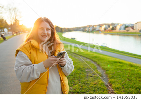 woman texting on phone in relaxed outdoor park near river with cotton and plastic case, casual and serene, yellow and green tones, frontal view, lifestyle and technology context. woman texting on phone in relaxed outdoor park near river with cotton and plastic case, casual and serene, yellow and green tones, frontal view, lifestyle and technology context. 141055035